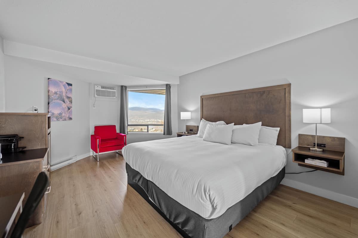 A modern hotel room with a large bed, a red chair, a desk, and a view of the mountains.