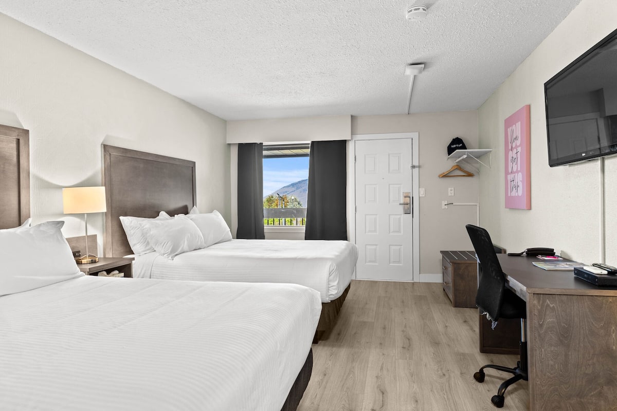 A hotel room with two beds, a desk, a chair, a TV, and a window with a view of a mountain.
