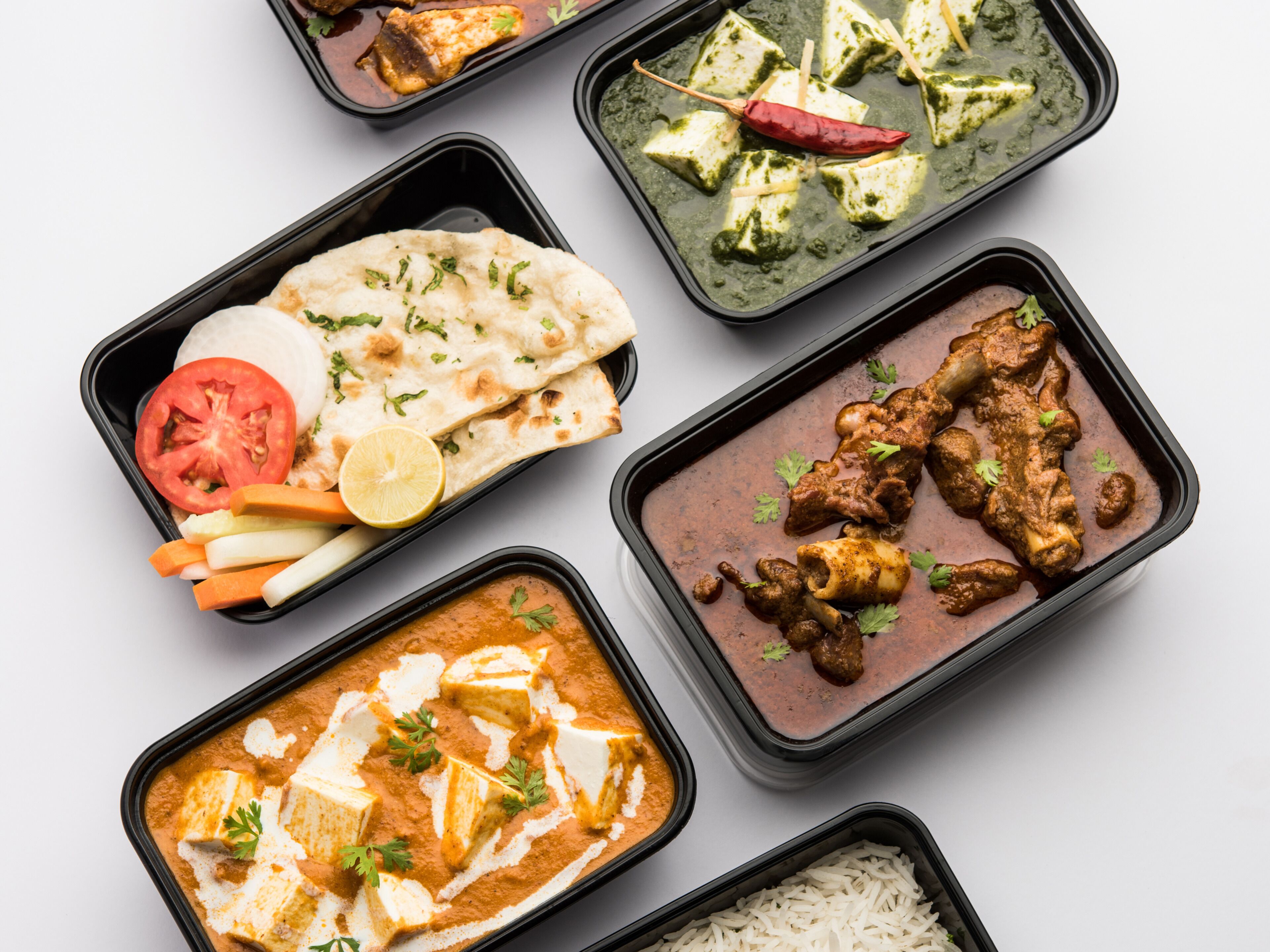 Variedad de platos de la cocina india en recipientes negros, incluyendo curries de paneer, pollo y verduras, acompañados de pan naan y arroz.