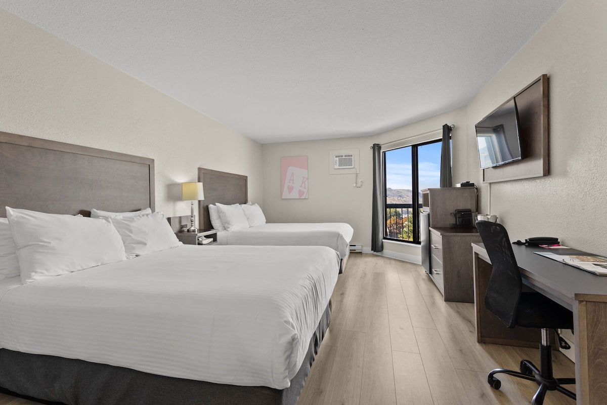 A hotel room with two beds, a desk, a chair, a TV, and a window with a view.