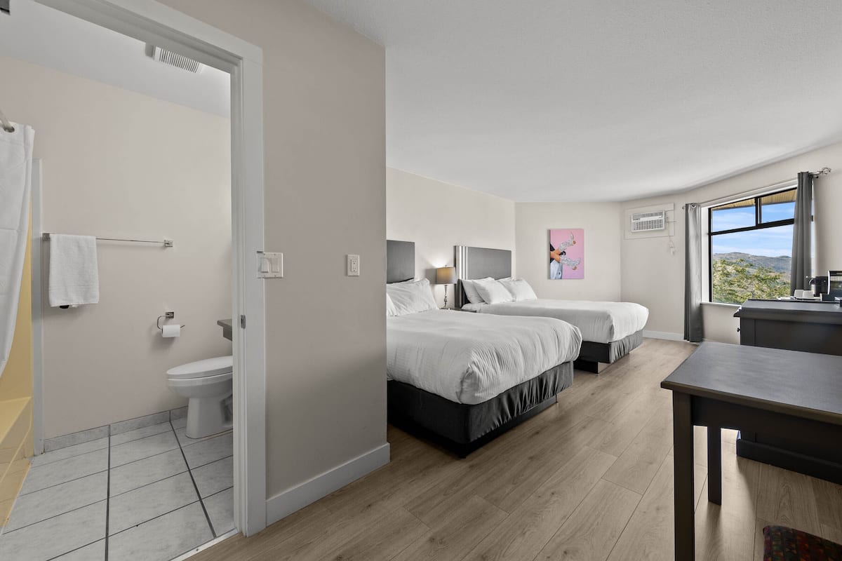 A modern hotel room with two beds, a bathroom, and a view of the outdoors.