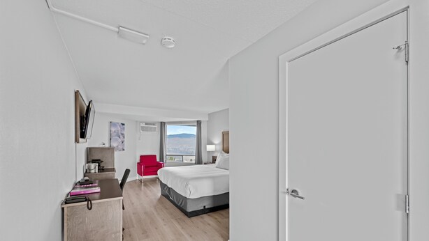 A modern hotel room with a bed, desk, and chair. There is a window with a view of the city. - The Vista Inn (Kamloops)