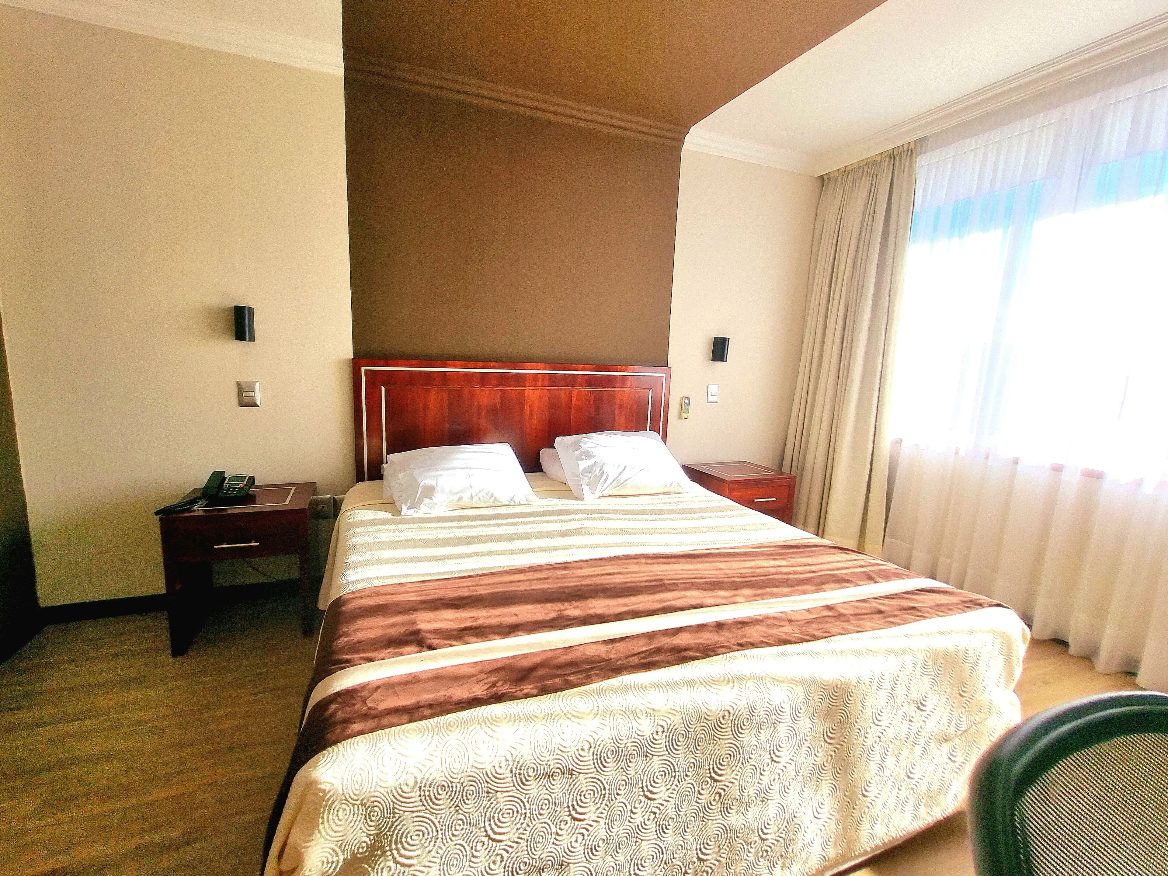 Une chambre d’hôtel avec un grand lit, une table de chevet avec un téléphone, une petite table de nuit et une fenêtre avec des rideaux.