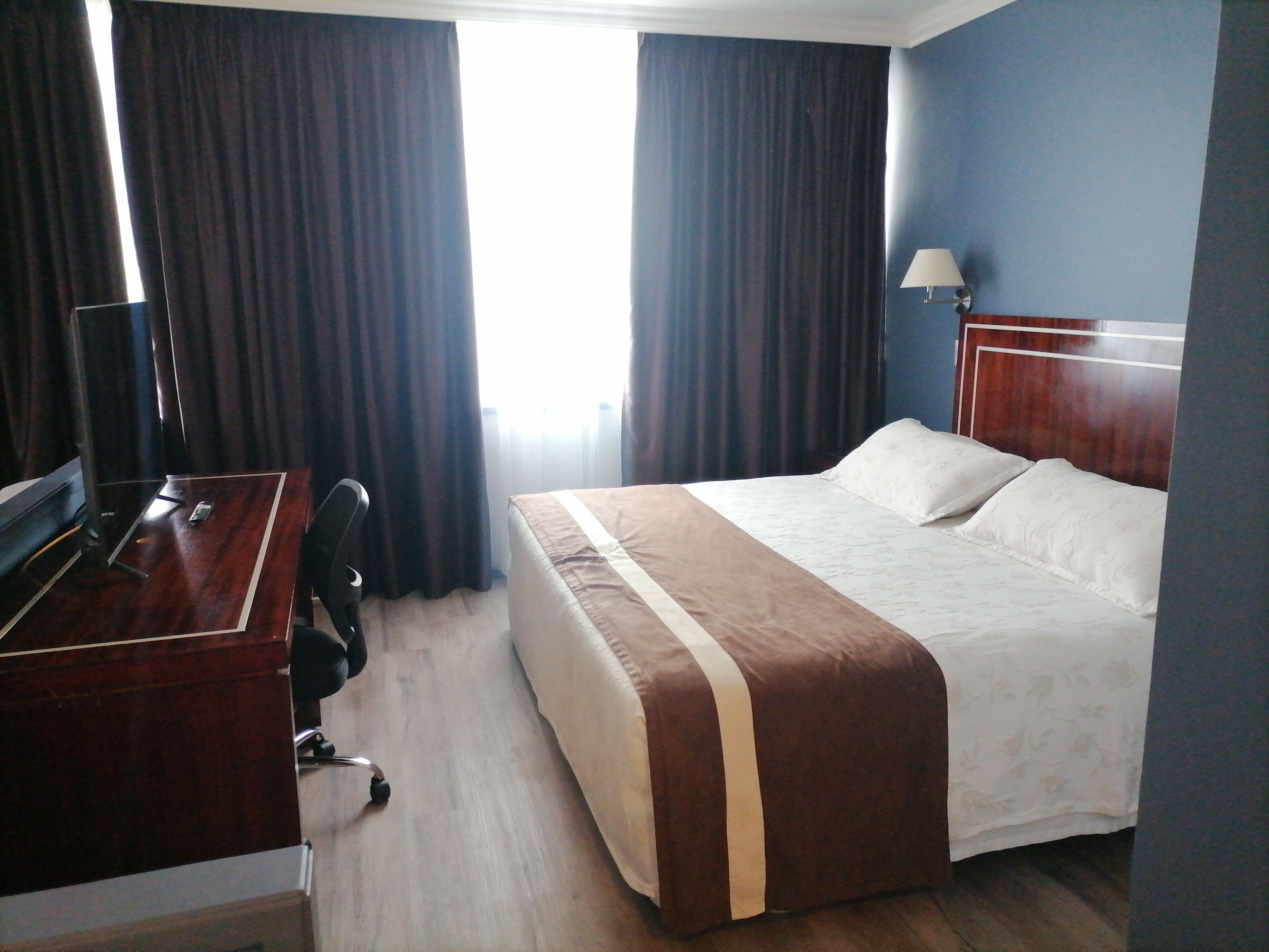A hotel room with a bed, a desk, a chair, a lamp, and a window with curtains.