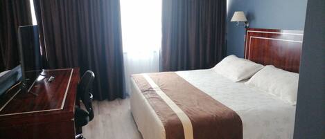 A hotel room with a bed, a desk, a chair, a lamp, and a window with curtains.