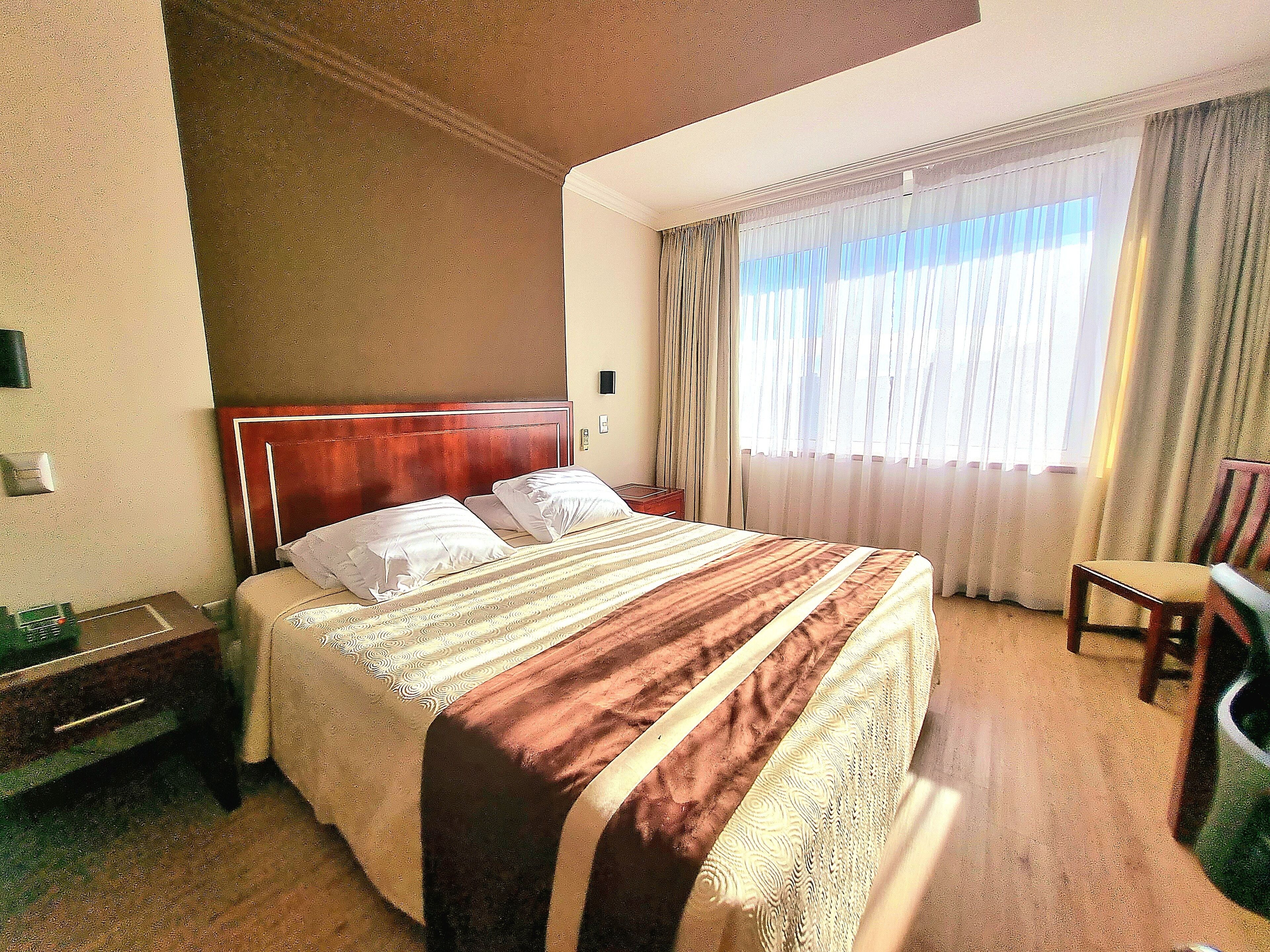 Habitación de hotel con una cama grande, cabecera de madera, mesita de noche, una silla y una ventana con cortinas.