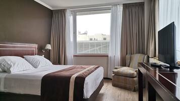 A hotel room with a bed, a desk with a TV, a chair, and a window with curtains.