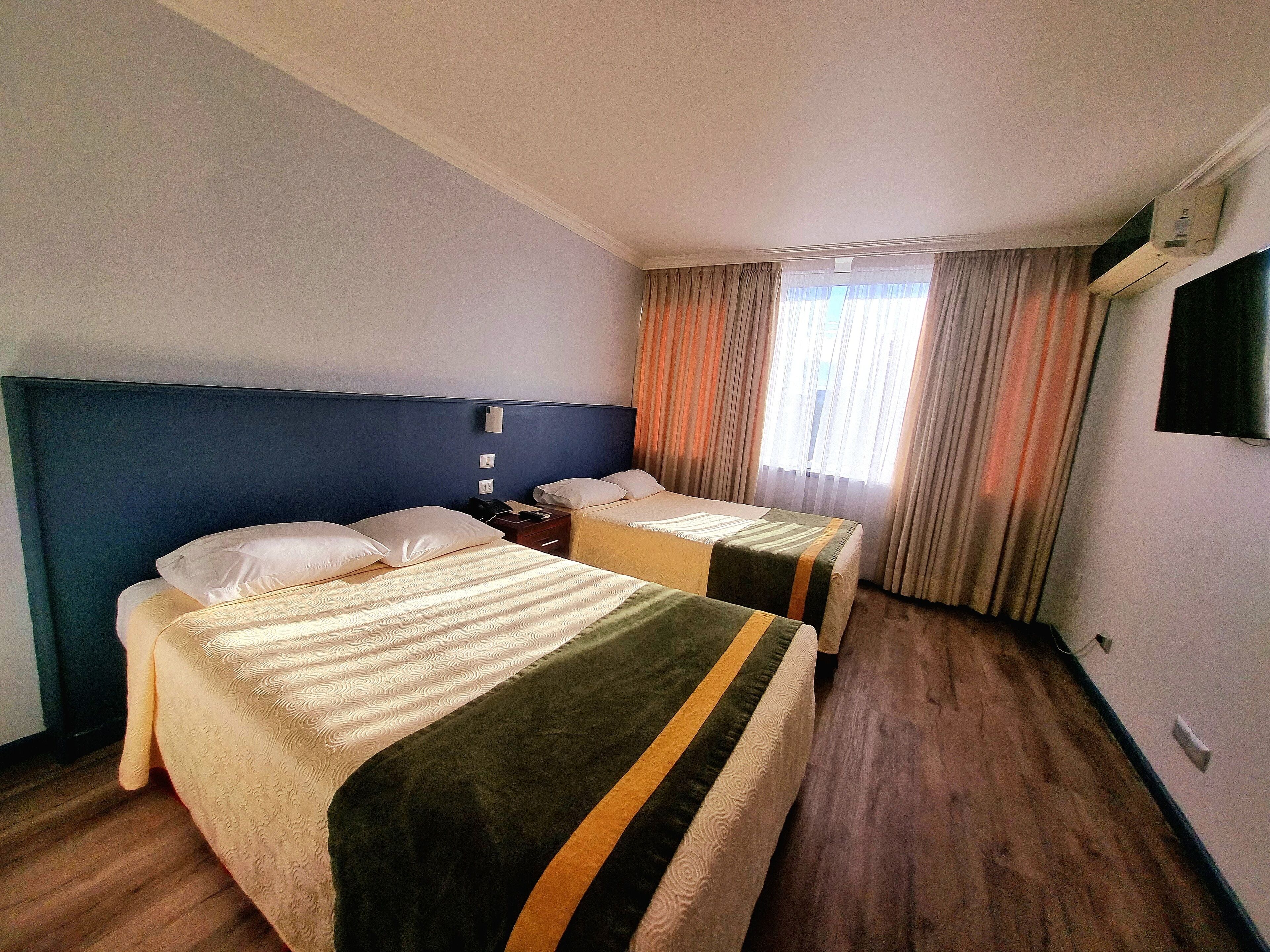 Une chambre d’hôtel avec deux lits, un téléviseur à écran plat, une table de chevet avec une lampe et une fenêtre avec des rideaux.