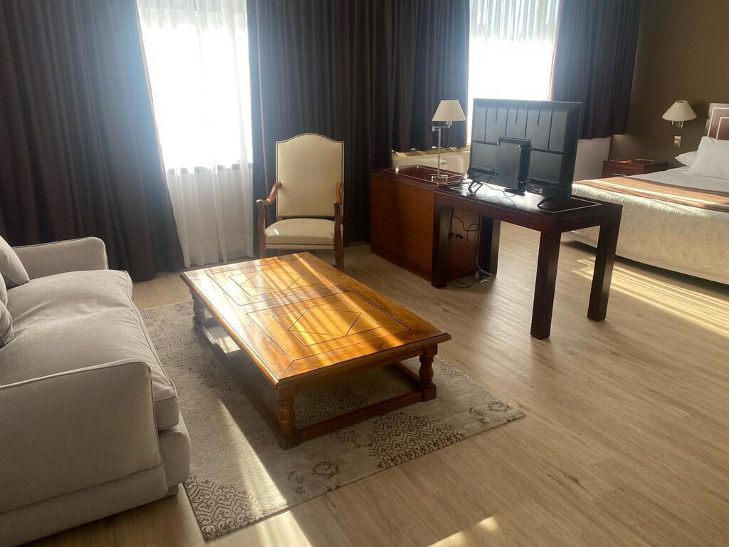 A hotel room with a sofa, a wooden coffee table, a desk with a computer, a TV, and a bed.