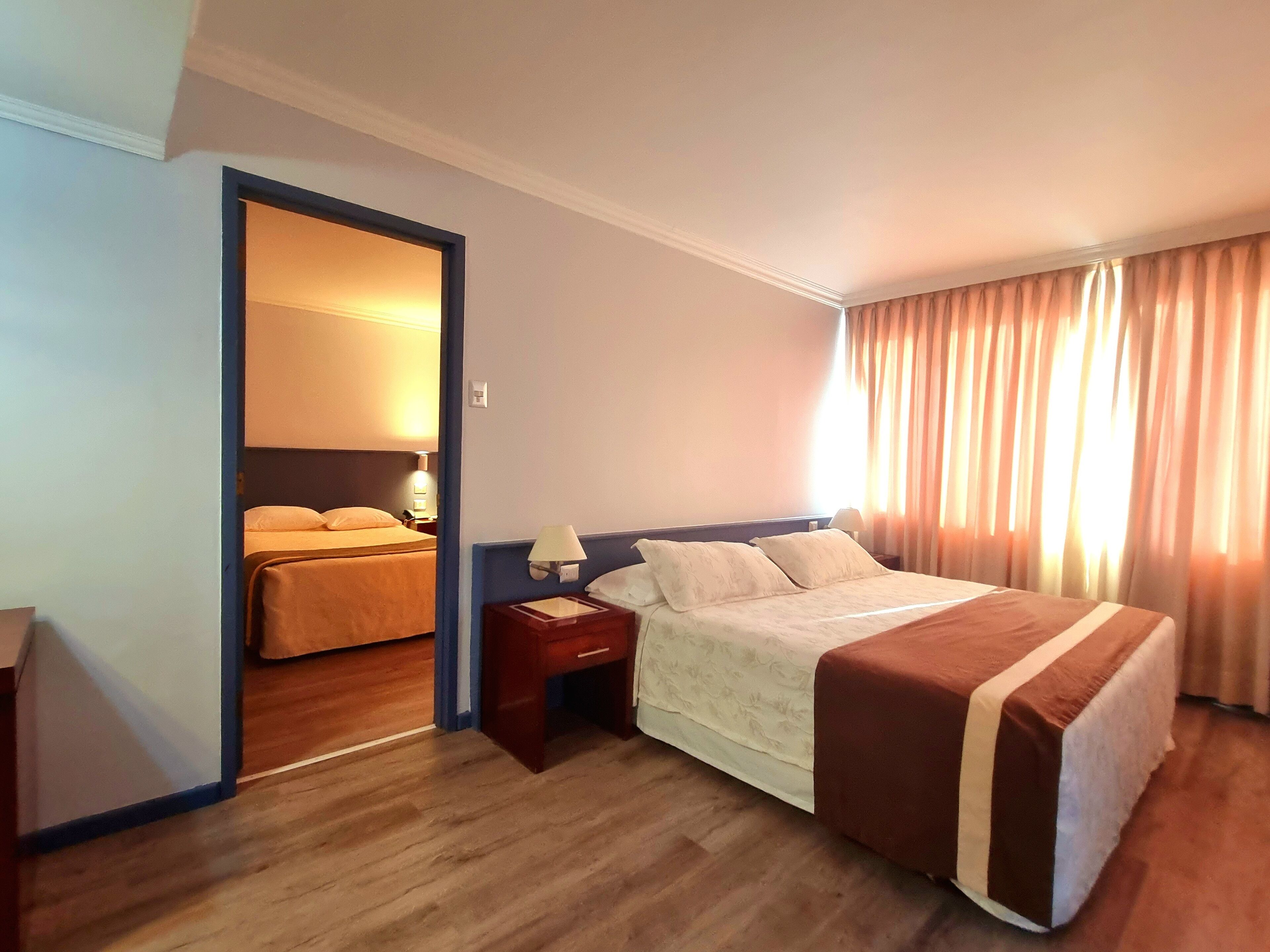 Habitación de hotel con una cama grande, una mesita de noche con lámpara, piso de madera y vista a otra habitación con una cama a través de una puerta.