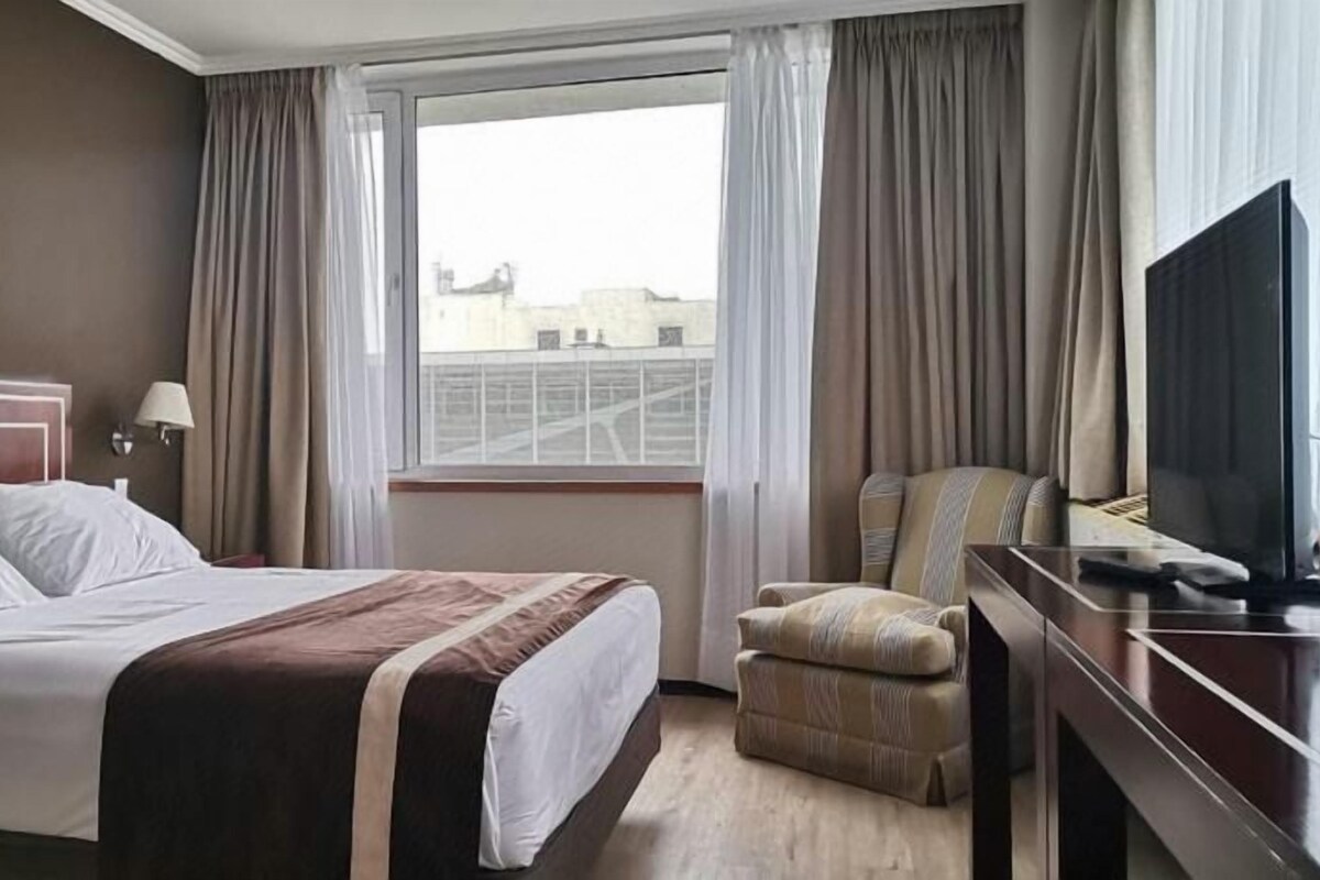 A hotel room with a bed, a desk with a television, a chair, and a window with curtains.