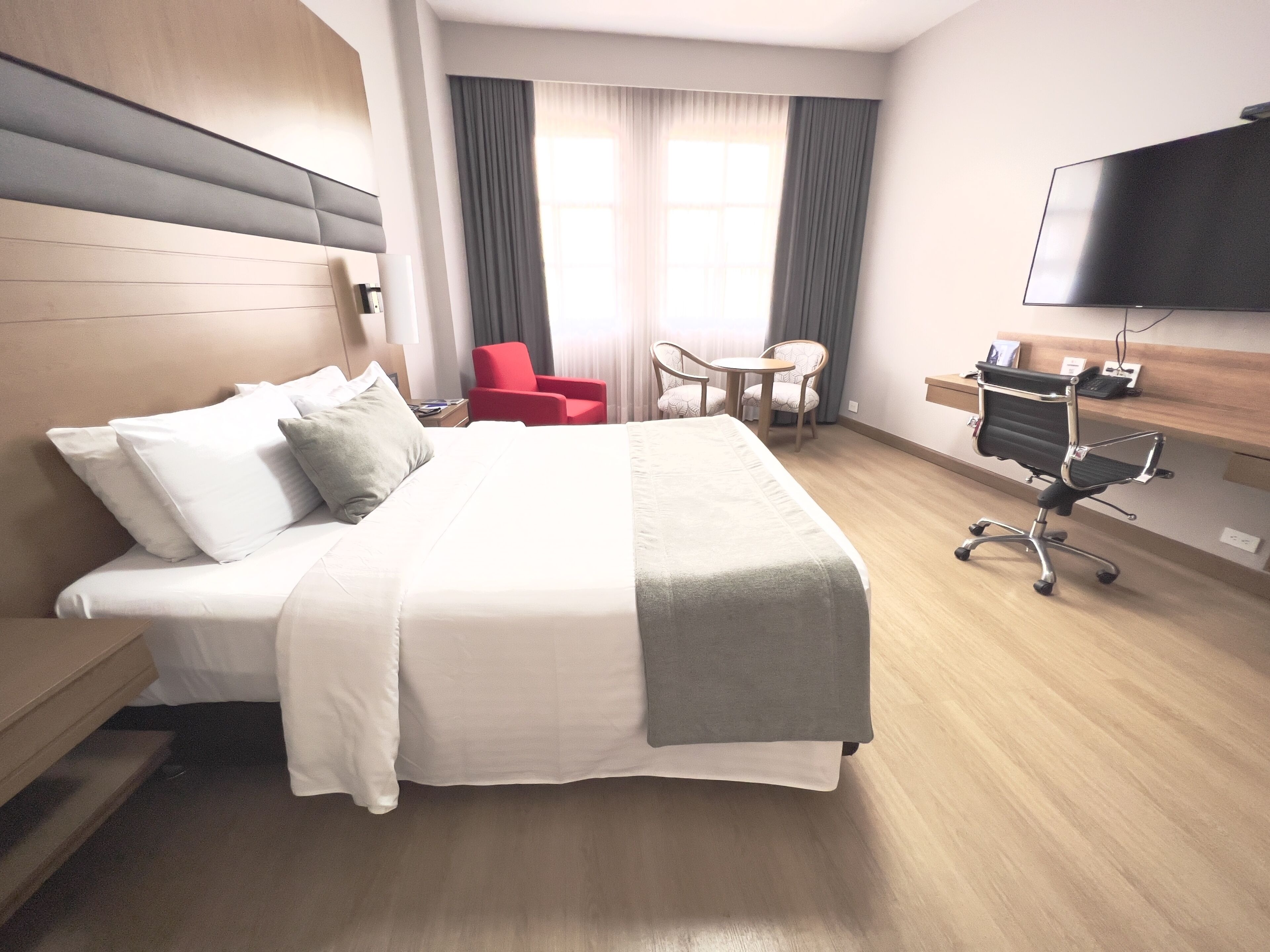 A hotel room with a large bed, a desk, a chair, a TV, and a window with curtains.
