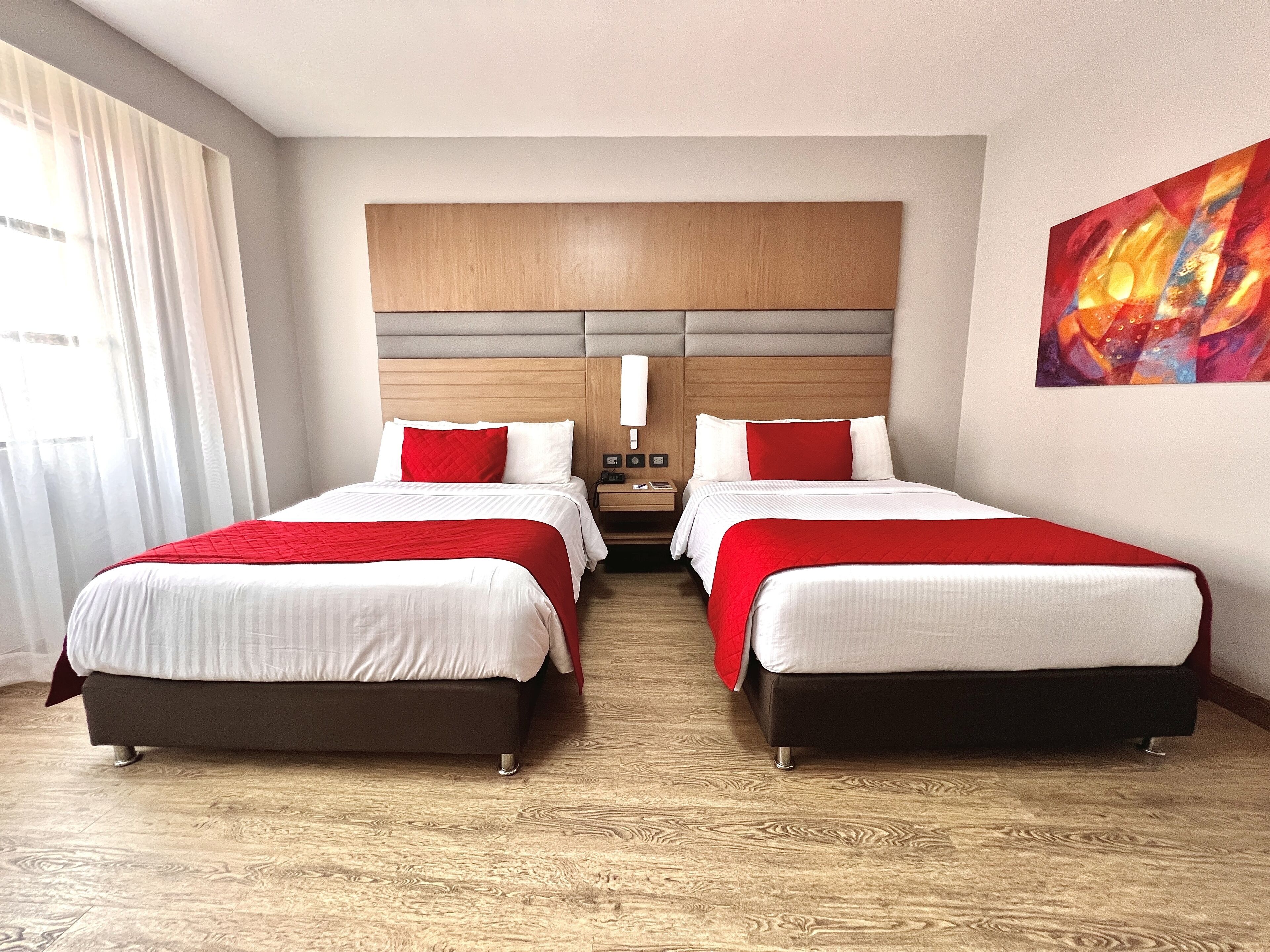Une chambre d’hôtel avec deux lits, une tête de lit en bois, un tableau au mur et une fenêtre avec des rideaux.