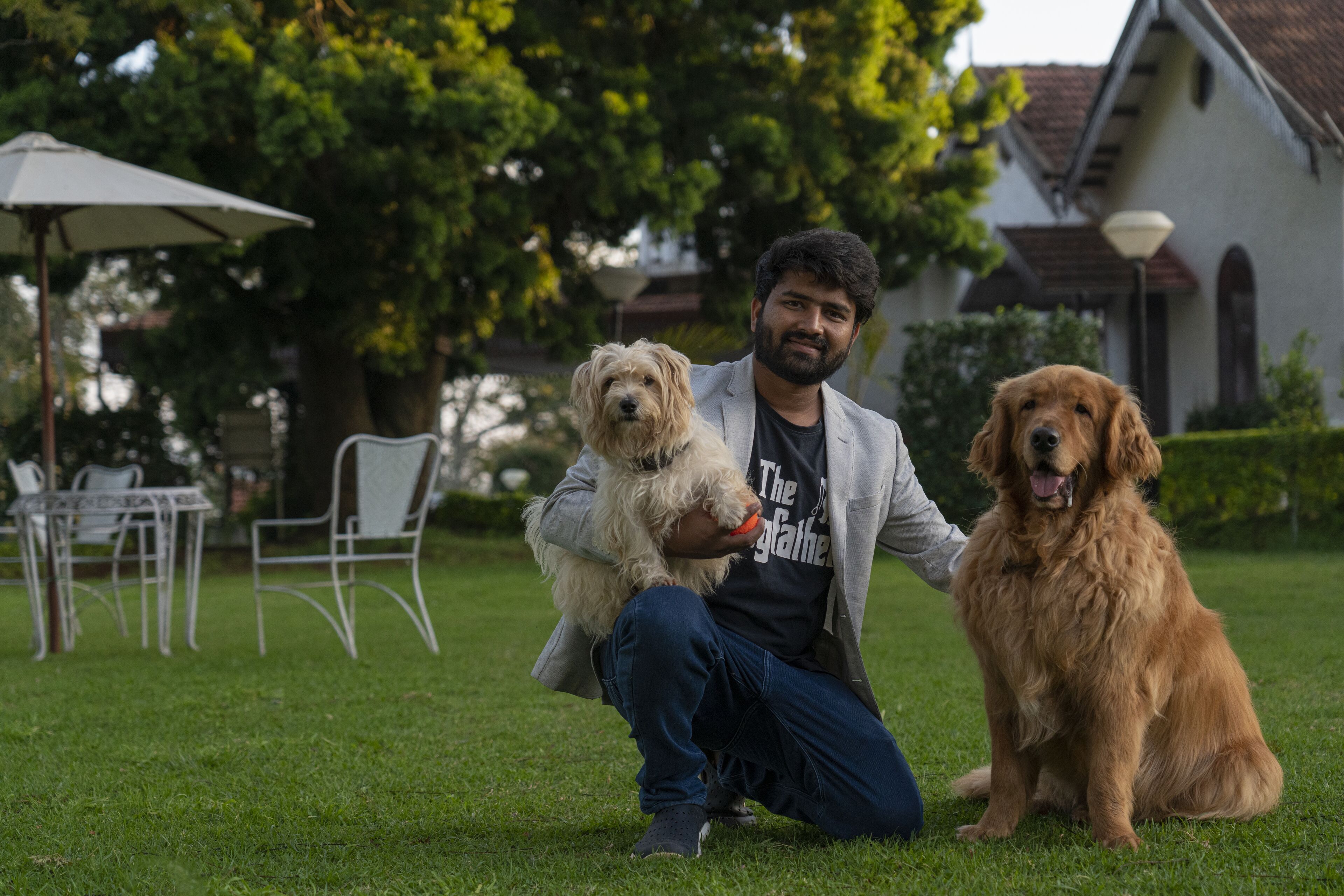 A person sitting on grass with two dogs, a white and a golden one, in a garden with a white building and a table with chairs in the background.