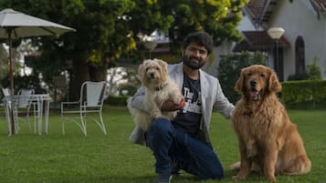 A person sitting on grass with two dogs, a white and a golden one, in a garden with a white building and a table with chairs in the background.