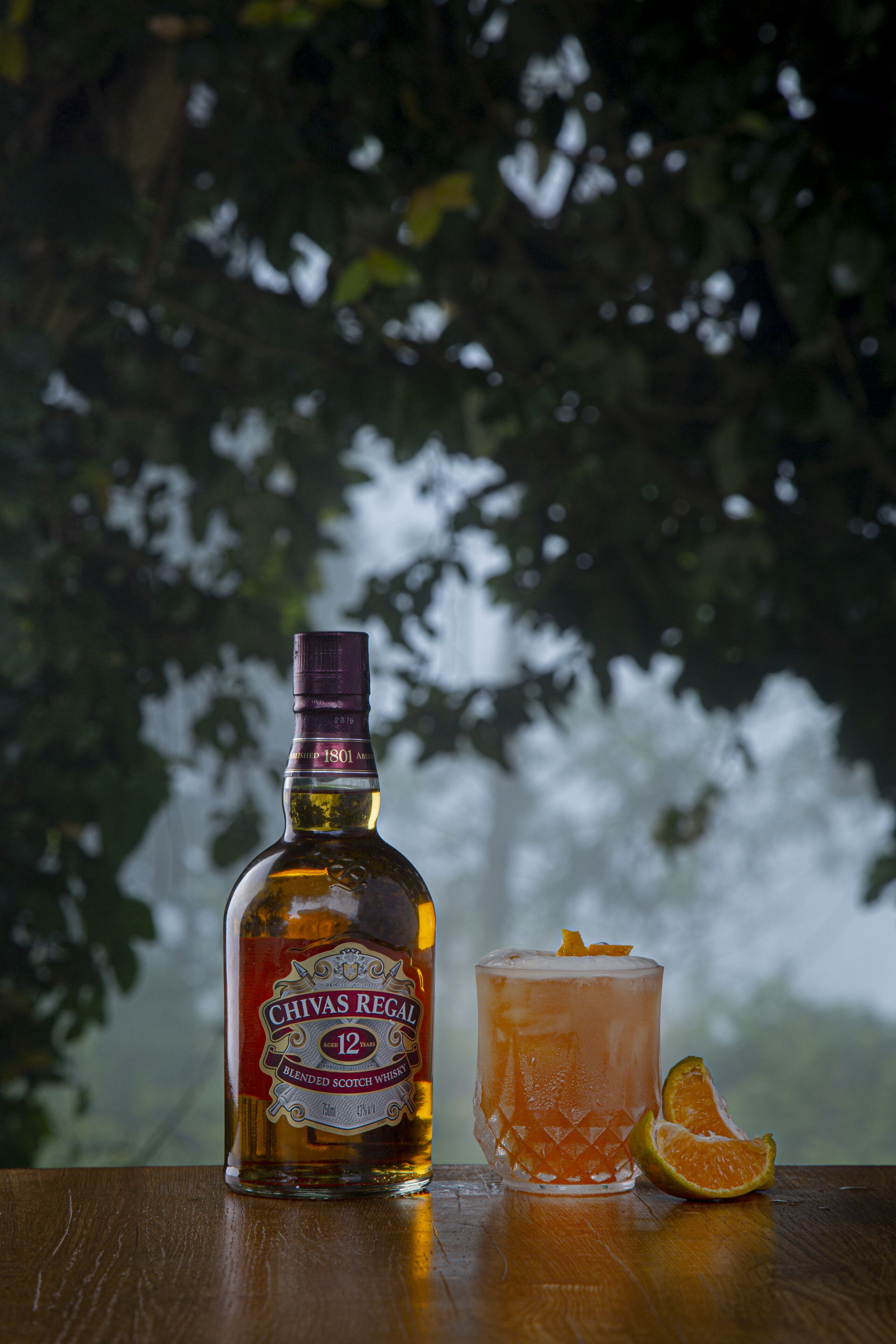 En flaska Chivas Regal 12, en blandad skotsk whisky, och ett glas med en apelsinskiva på en träyta.
