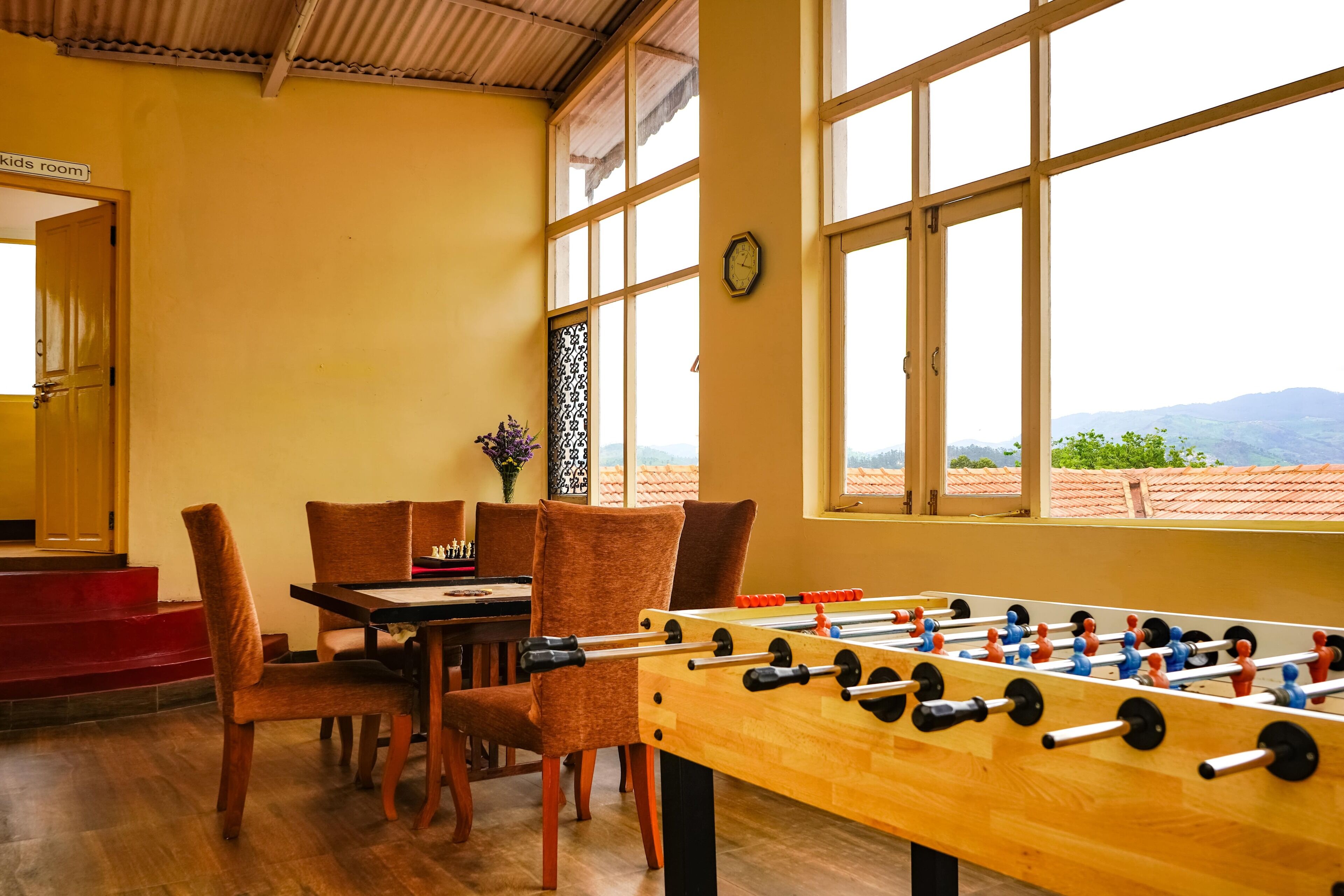 A room with a foosball table, wooden chairs, a dining table, and a window with a view of mountains.