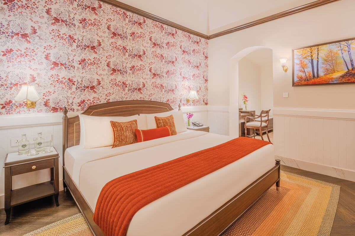 A hotel room with a large bed, a nightstand with a lamp, a desk with a chair, and a painting on the wall.