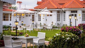 A white building with a red-tiled roof, outdoor seating with white wicker chairs and tables, and a well-maintained garden with pink flowers. - Gateway Coonoor (Coonoor)