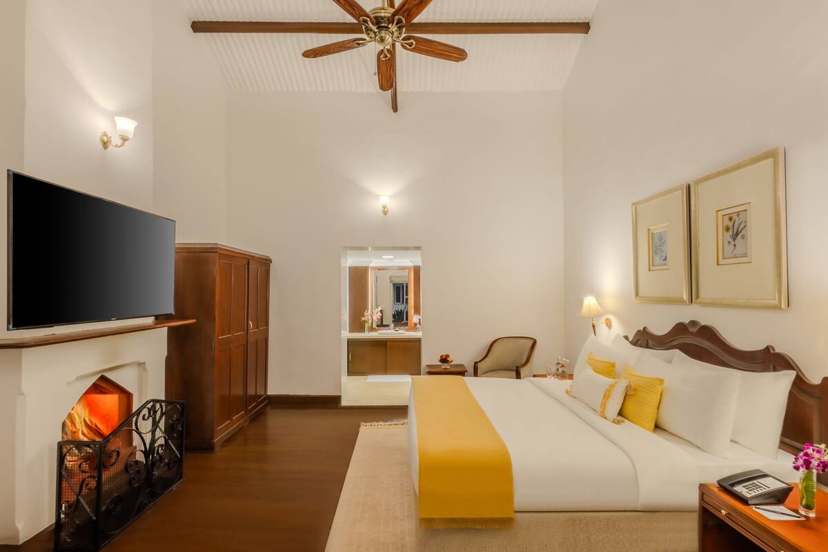A hotel room with a bed, a television, a fireplace, a ceiling fan, and framed artwork on the wall.