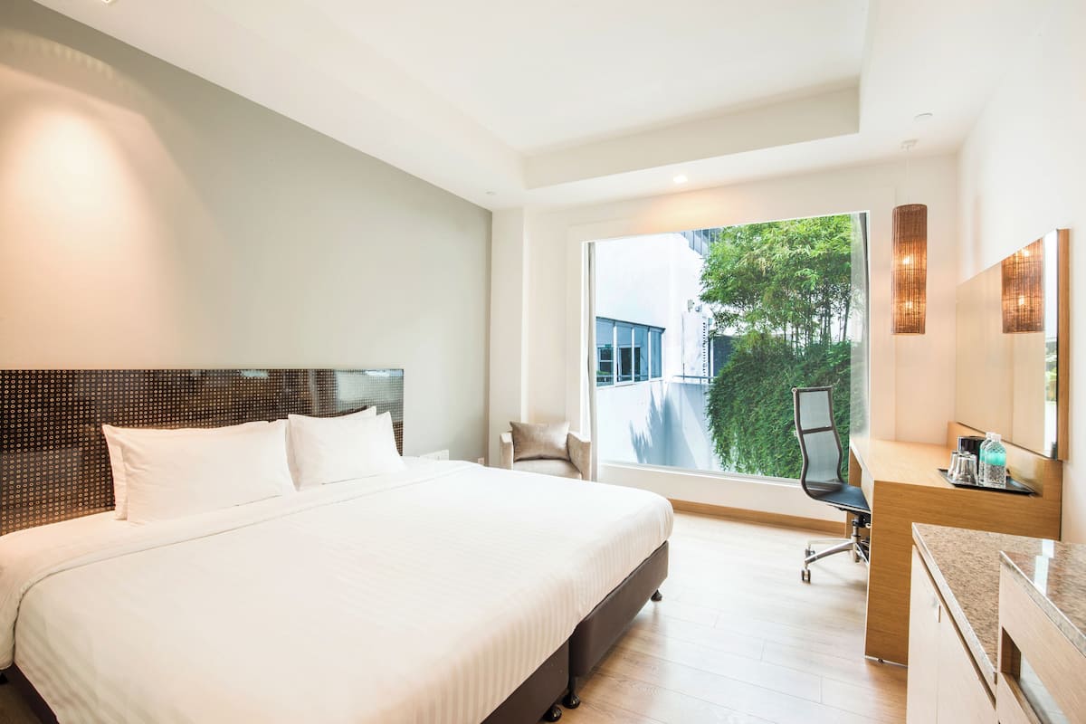 A modern hotel room with a large bed, a desk, and a chair. There is a window with a view of greenery outside.