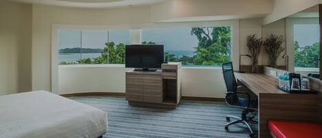Habitación de hotel con cama, escritorio con televisor, silla, banco rojo y ventana grande con vista a árboles y agua.