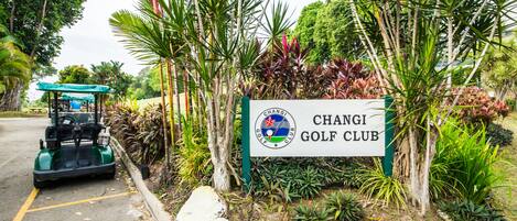 Señal de club de golf con un logotipo, rodeada de plantas tropicales y un carrito de golf.