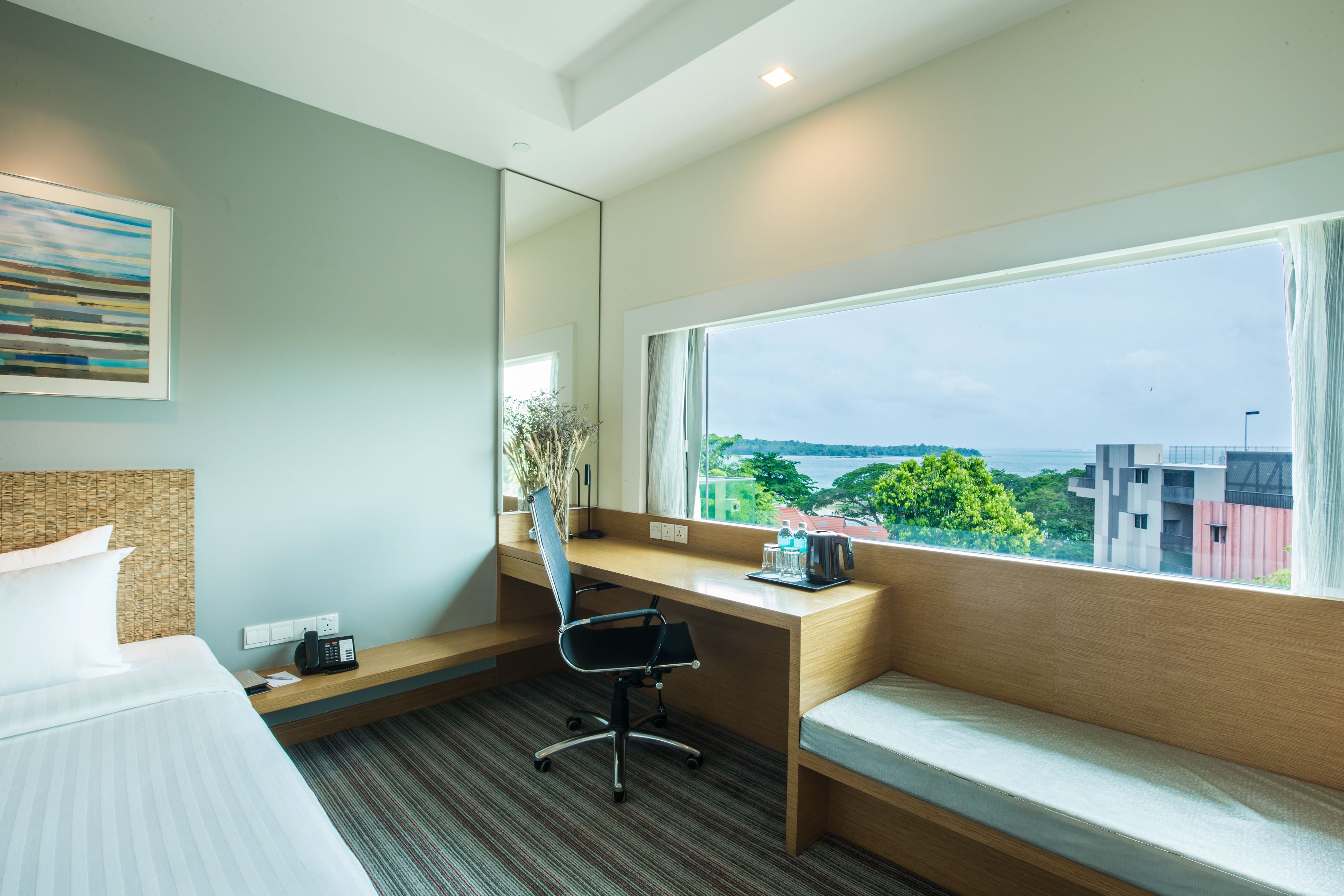 Habitación de hotel con cama, escritorio, silla, ventana grande con vistas a edificios y árboles, y un cuadro en la pared.