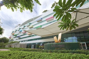 A modern hotel with a multi-story structure, balconies with colorful railings, and a prominent entrance with a green hedge.