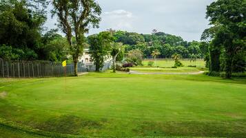 A well-maintained golf course with a fenced area, trees, and a distant view of a hill with buildings.
