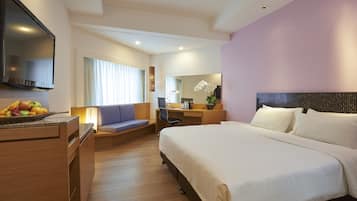 Ein modernes Hotelzimmer mit einem großen Bett, einem Schreibtisch mit Computer, einem Flachbildfernseher und einer Obstschale auf dem Nachttisch.