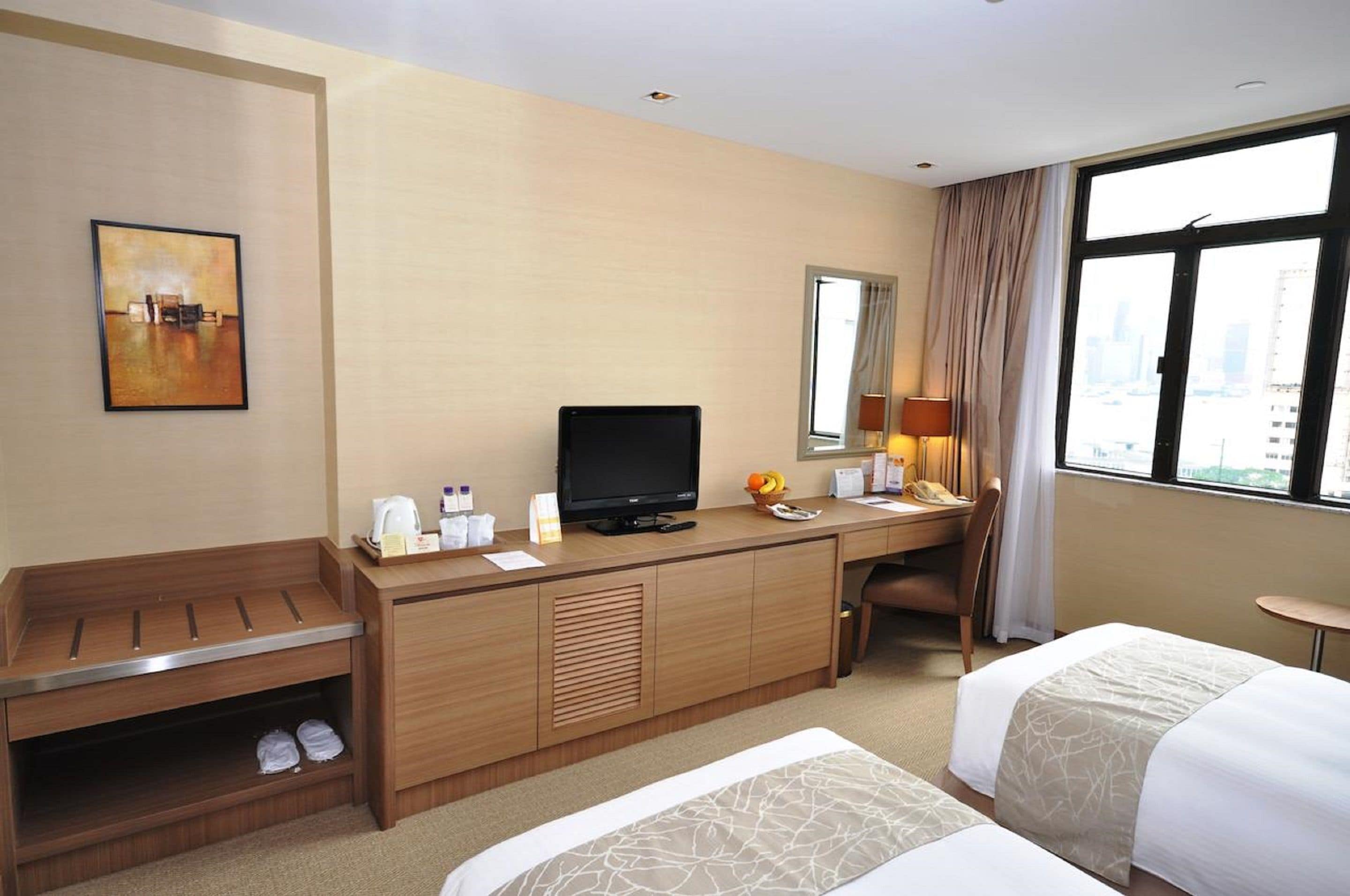 A hotel room with a bed, a desk with a television, a chair, a window with a city view, and a painting on the wall.