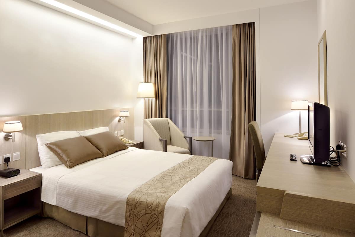 Hotellihuone, jossa on sänky, yöpöydällä olevat lamput, peilillä varustettu työpöytä, tuoli ja suuri ikkuna, jossa on verhot.