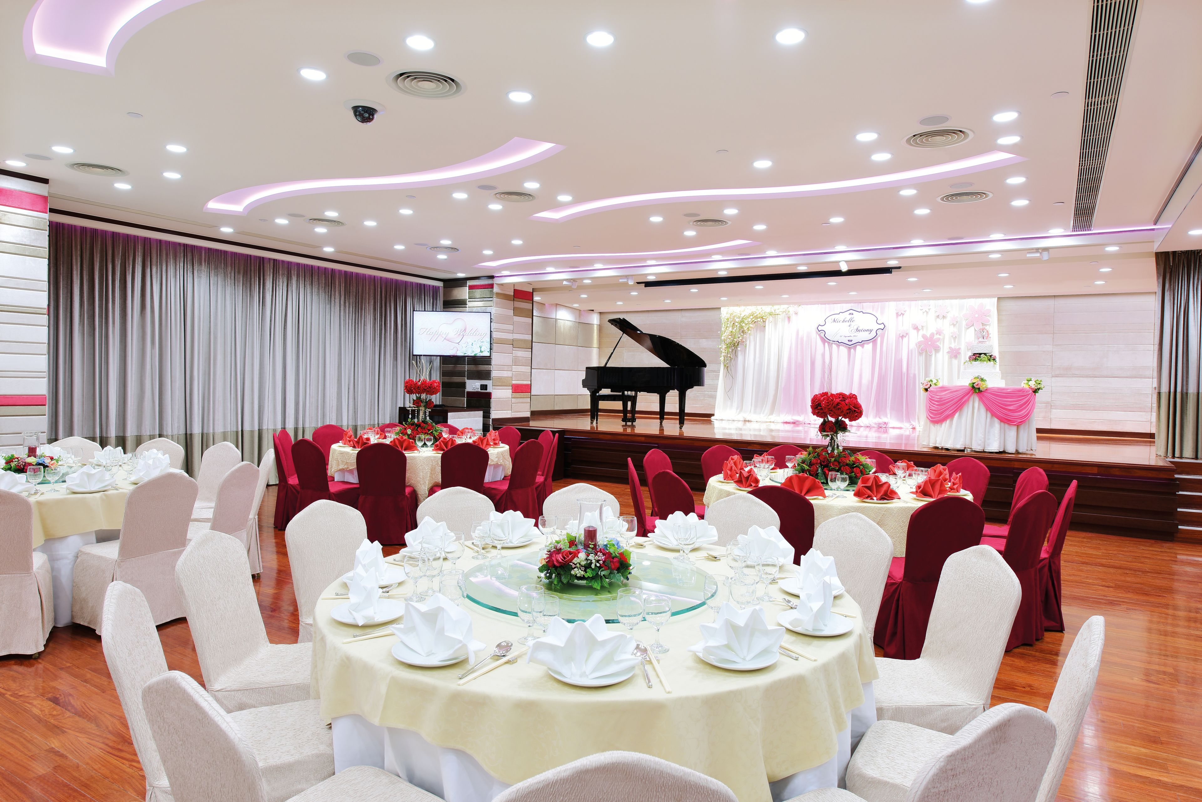A banquet hall with round tables set for a formal event, a stage with a grand piano, and a large screen.