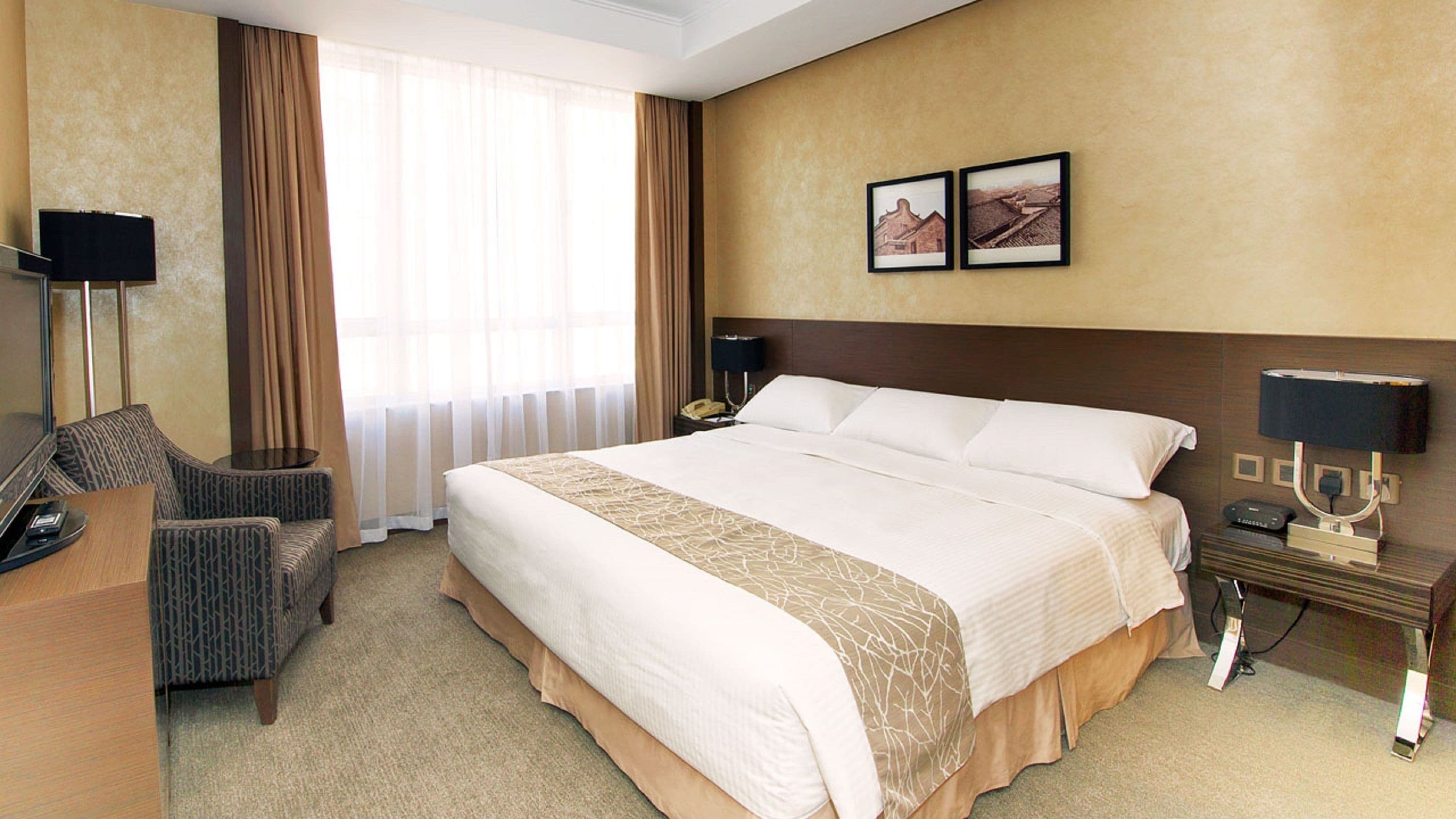 A hotel room with a large bed, a desk, a chair, a nightstand, a lamp, and two framed pictures on the wall.