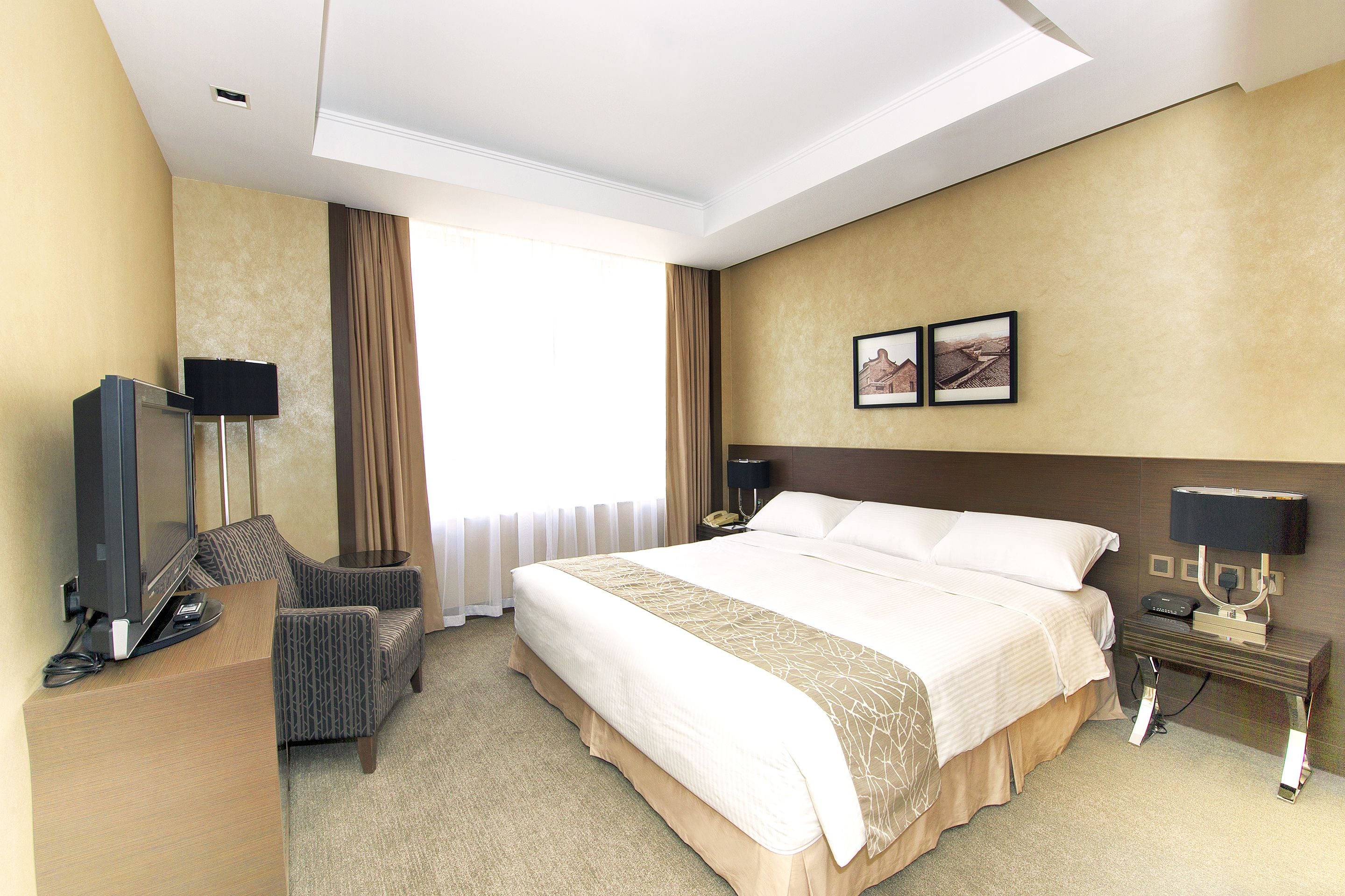 A hotel room with a large bed, a desk with a television, a chair, and a nightstand with a lamp.
