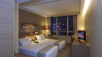 Habitación de hotel con dos camas, un escritorio con televisor, una ventana con vista a la ciudad y un cuadro en la pared.