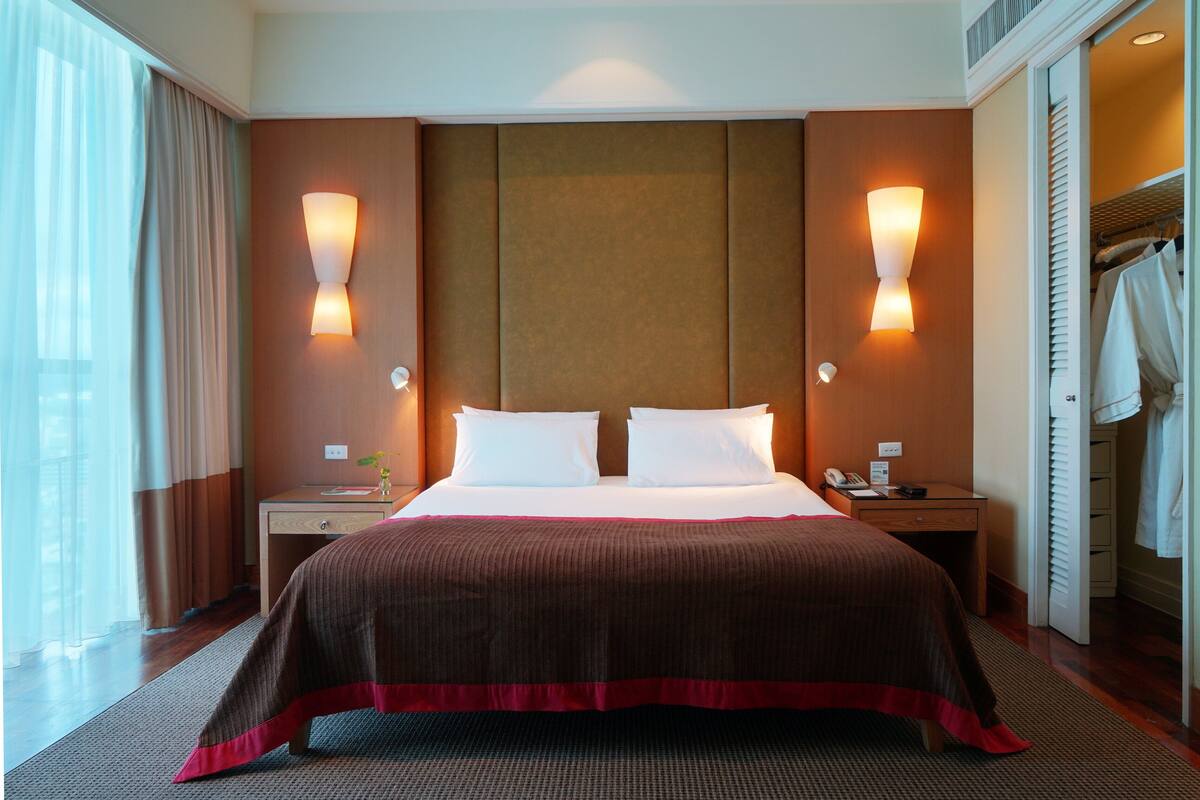 A hotel room with a large bed, two bedside tables, a nightstand, a wardrobe, and a window with curtains.