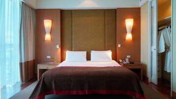 A hotel room with a large bed, two bedside tables, a nightstand, a mirror, a window with curtains, and a wardrobe.