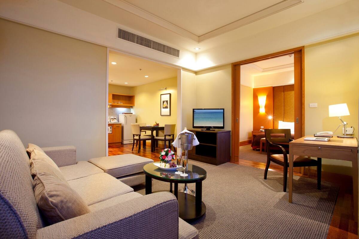A hotel room with a sofa, a dining table, a TV, and a desk with a lamp.