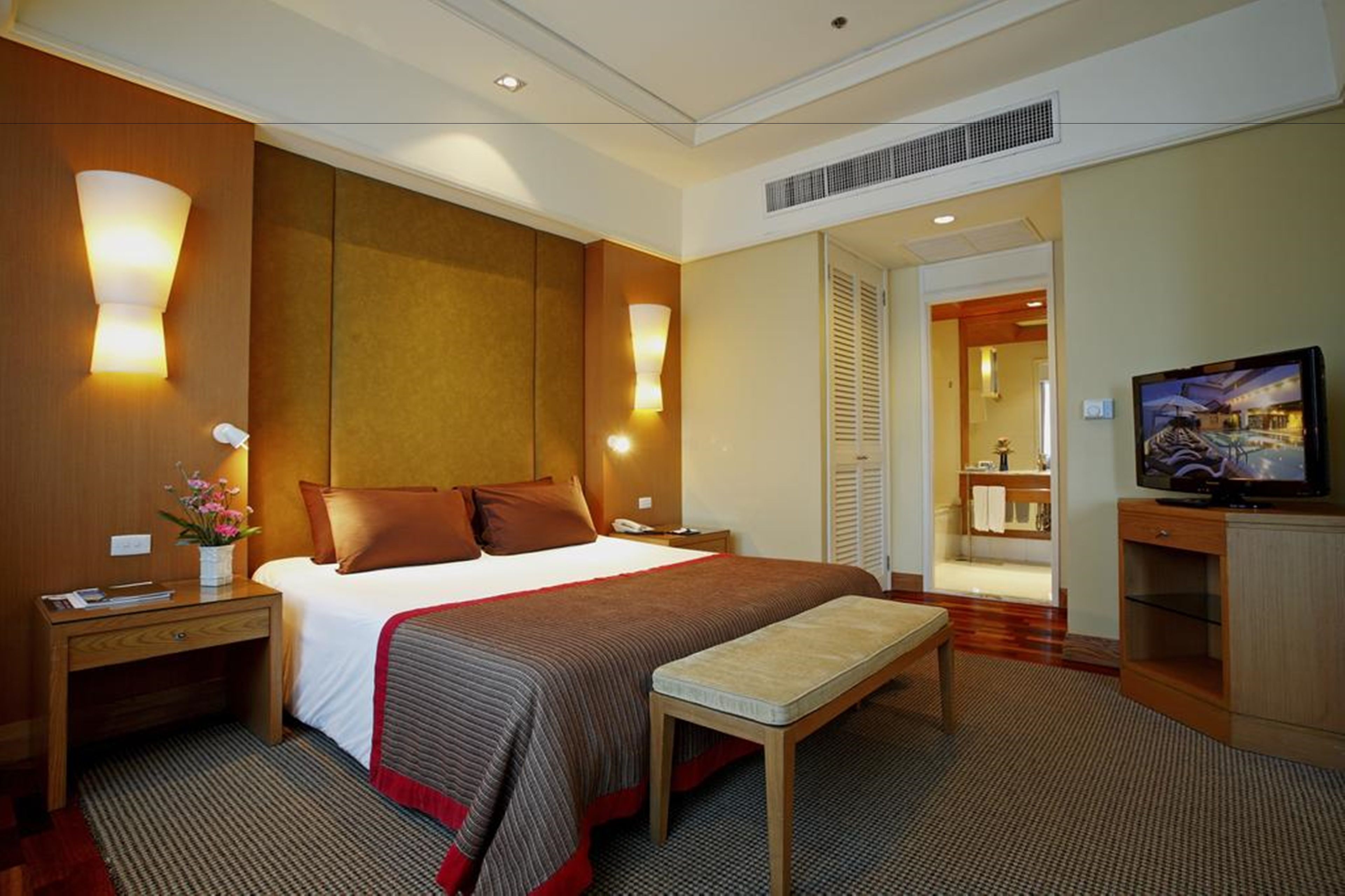 A hotel room with a large bed, bedside tables, a bench, a television, and a bathroom visible through an open door.