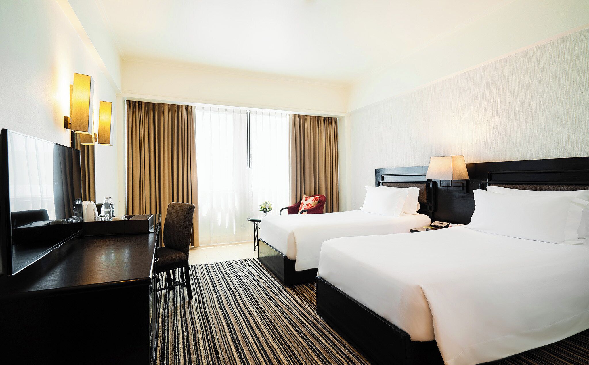 A hotel room with two beds, a desk, a TV, and a large window with curtains.