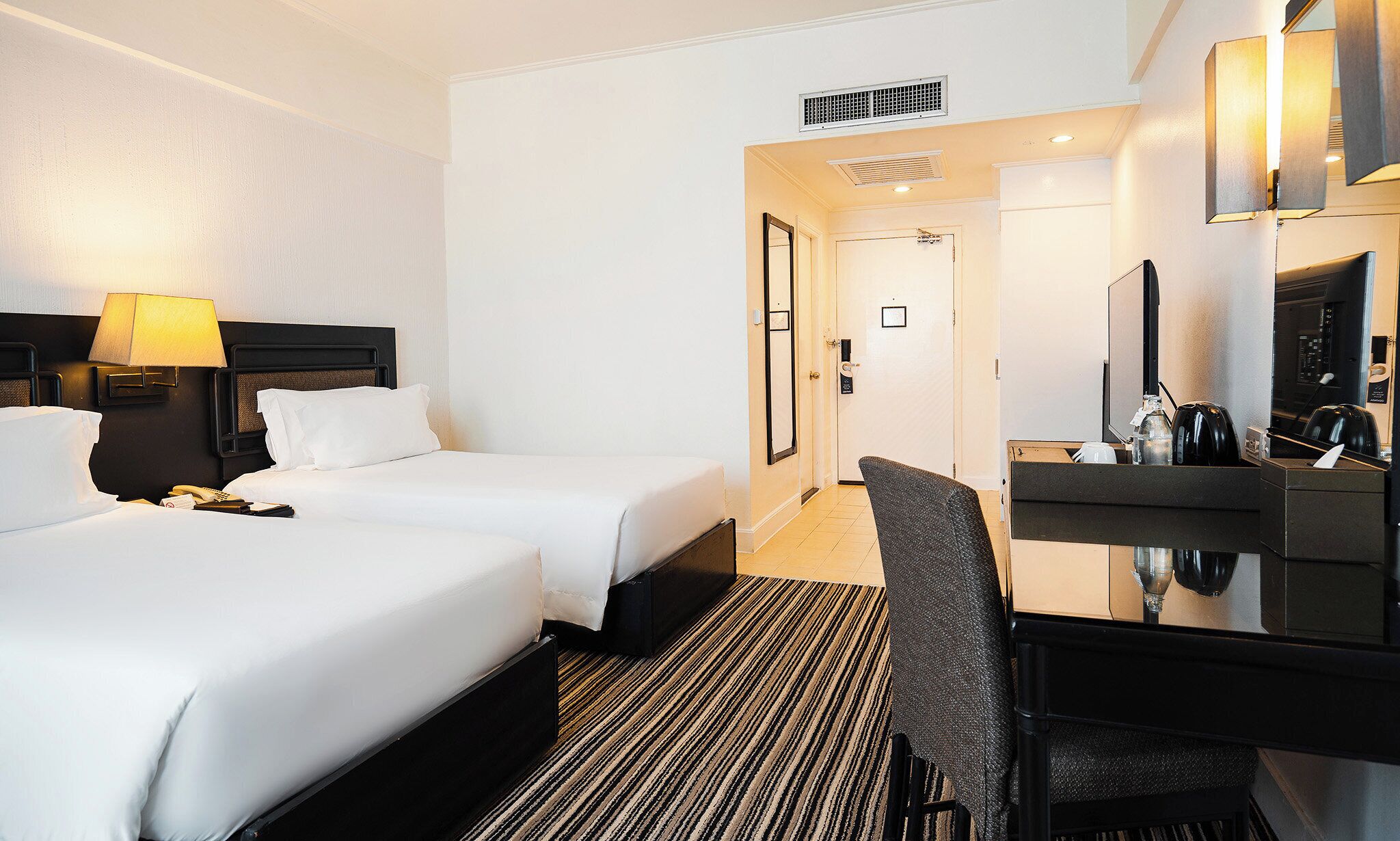 Habitación de hotel con dos camas, un escritorio con silla, un espejo y una zona de kitchenette.