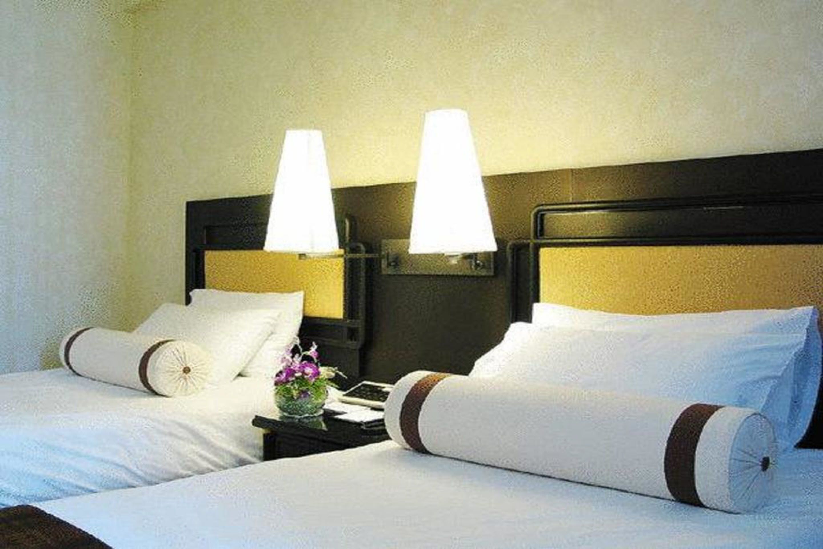 A hotel room with two beds, each with white linens and brown trim, a nightstand with a lamp, and a small vase with flowers.
