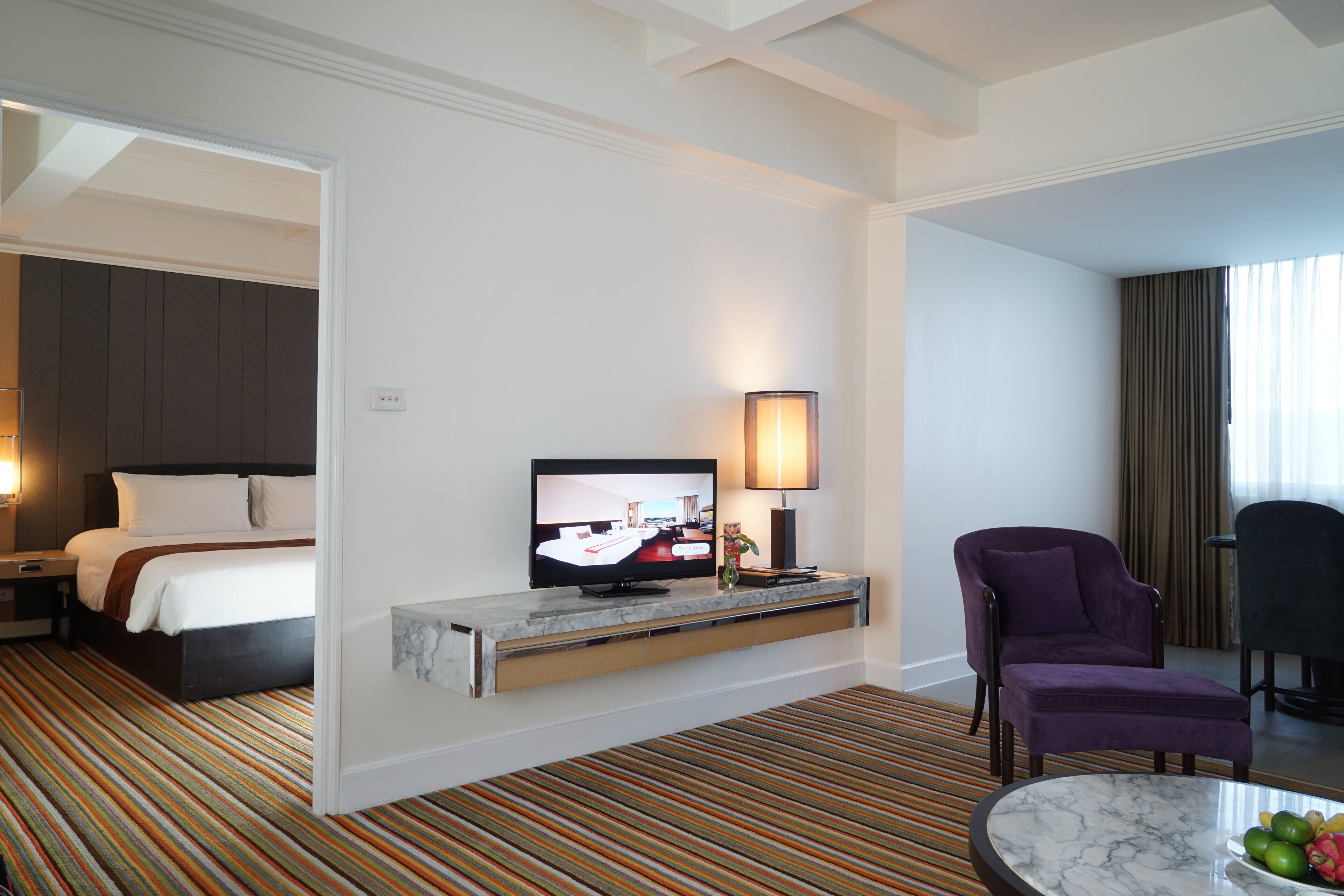 Habitación de hotel con una cama, un televisor en un soporte, un sillón púrpura, una mesita con fruta y una alfombra a rayas.