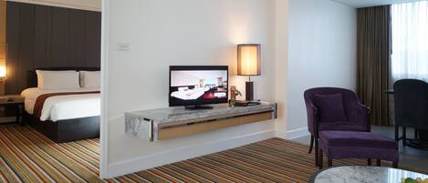 Habitación de hotel con una cama, un televisor en un soporte, un sillón púrpura, una mesita con fruta y una alfombra a rayas.