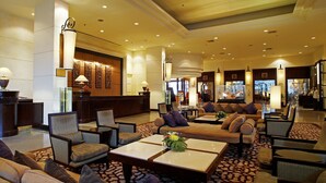 Eine großzügige Lobby mit mehreren Sitzbereichen, darunter Sofas und Sessel, die um einen zentralen Couchtisch angeordnet sind. Im Hintergrund sind dekorative Wandpaneele und ein Fernsehbild sichtbar.