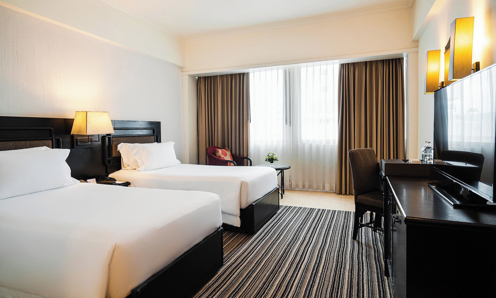 A hotel room with two beds, a desk with a computer, a chair, a lamp, and a large window with curtains.