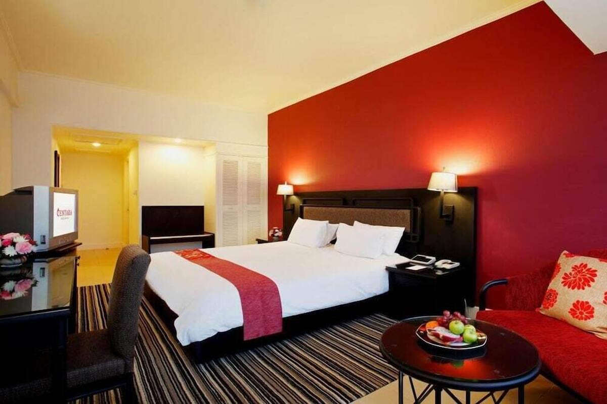 A hotel room with a bed, bedside tables, a desk with a lamp, a TV, and a red wall.
