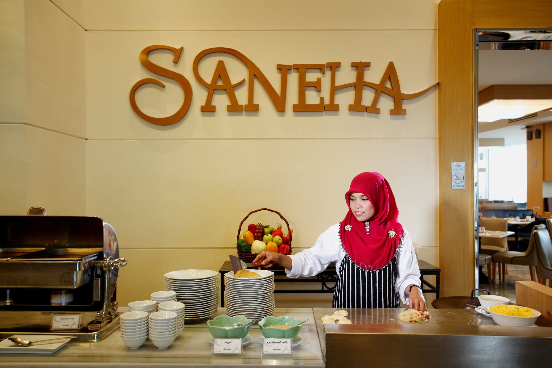 Montaje de bufé con un letrero que dice "SANEHA" en la parte superior, una barra de bufé con platos y recipientes, y un miembro del personal sirviendo comida.